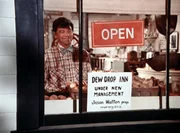 Ike (Joe Conley) stellt ein Plakat in das Schaufenster, auf dem die Neuer&ouml;ffnung des Dew Drop Inn angek&uuml;ndigt wird. Jason hatte das Lokal gekauft und will es nun selbst betreiben.