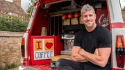 ant anstead, wearing black tshirt and jeans, sitting with arms crossed in the back of the ford transit van AFTER it has been converted to a coffee truck, with machine and coffee cups and sign in view, parked infront of the farm brick wall