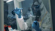 Ein Labortechniker schüttelt ein Reagenzglas hinter einer Schutzscheibe in einem BSL-Labor. Ein Labortechniker schüttelt ein Reagenzglas hinter einer Schutzscheibe in einem BSL-Labor.