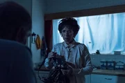 Bev (ADRIENNE PIERCE) enters the kitchen and finds Doug (DEON LOTZ) sitting at the kitchen table shrouded in the darkness.