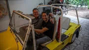 Mike and Elvis seated in yellow Yellow Mini Moke looking into stills camera smiling
