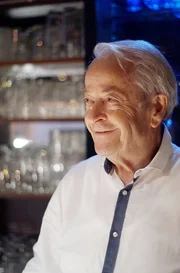 Ein alter Mann mit wei&szlig;em Hemd steht in einer dunklen Gastst&auml;tte vor einem Gl&auml;serregal und l&auml;chelt. Achim Wenning ist 82 und immer noch Gastwirt mit Leib und Seele.