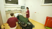 Berlin: two members of staff are around a laying patient. The male one is sitting on a white stool, the female one is standing at the head of the bed seemingly bringing the patient something to drink. To the right we see a big trash container with a red bag. Berlin: two members of staff are around a laying patient. The male one is sitting on a white stool, the female one is standing at the head of the bed seemingly bringing the patient something to drink. To the right we see a big trash container with a red bag.