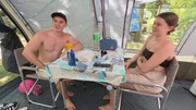 This image shows two people sitting at a small folding table inside a camping tent setup on a sunny day. This image shows two people sitting at a small folding table inside a camping tent setup on a sunny day.