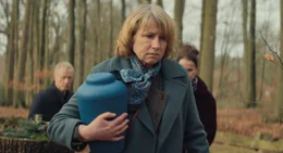 Lissy Lunies (Corinna Harfouch) tr&auml;gt bei einer Beisetzung die Urne mit der Asche ihres Mannes durch einen Wald.