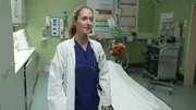 Lübeck: Neurologin Valentina gibt scheinbar ein Interview. Sie trägt einen weißen Kittel über blauen OP-Kleidern und befindet sich im Schockraum. Im Hintergrund ist ein leeres Bett mit weißem Laken zu sehen; der Raum ist grün gefliest. Links stehen hohe Schränke, im Hintergrund unter anderem medizinische Geräte und ein Rollwagen. Lübeck: Neurologin Valentina gibt scheinbar ein Interview. Sie trägt einen weißen Kittel über blauen OP-Kleidern und befindet sich im Schockraum. Im Hintergrund ist ein leeres Bett mit weißem Laken zu sehen; der Raum ist grün gefliest. Links stehen hohe Schränke, im Hintergrund unter anderem medizinische Geräte und ein Rollwagen.