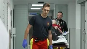 Herne: dynamic shot of two paramedics moving a gurney on the aisle. The one in the front is wearing a black T-shirt, red-yellow trousers and blue gloves and looking to the side. On the left we see the back of a physician in his white coat. Herne: dynamic shot of two paramedics moving a gurney on the aisle. The one in the front is wearing a black T-shirt, red-yellow trousers and blue gloves and looking to the side. On the left we see the back of a physician in his white coat.