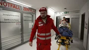 Füssen: dynamic shot of a bearded paramedic wearing all red, a cap and glasses pulling a gurney with a new pixelated patient into the A&E. To his right we see in big red letters the word for A&E Center in German over glass doors. Behind him and somewhat blurry we see a laying patient and a colleague of him. Füssen: dynamic shot of a bearded paramedic wearing all red, a cap and glasses pulling a gurney with a new pixelated patient into the A&E. To his right we see in big red letters the word for A&E Center in German over glass doors. Behind him and somewhat blurry we see a laying patient and a colleague of him.